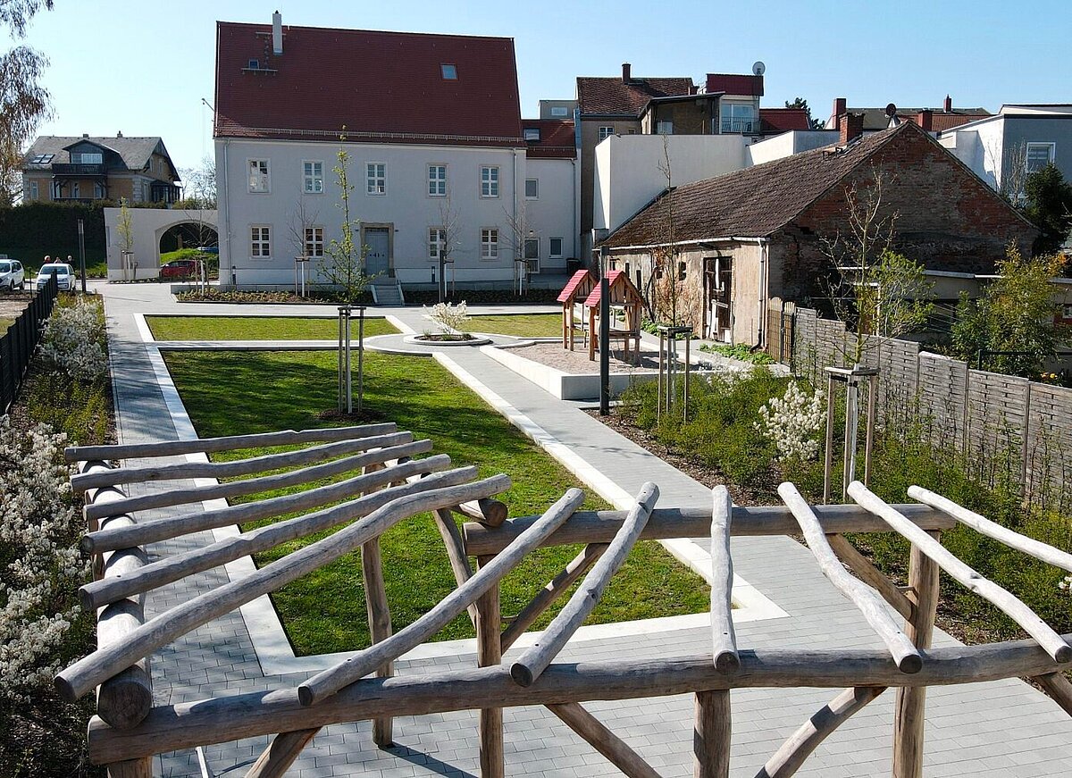 Parkanlage mit Wegen, Pflanzflächen und Spielbereich vor historischen Gebäuden