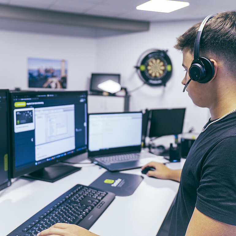 Azubi arbeitet mit Headset an mehreren Monitoren an einem Computerarbeitsplatz bei Dataflor.